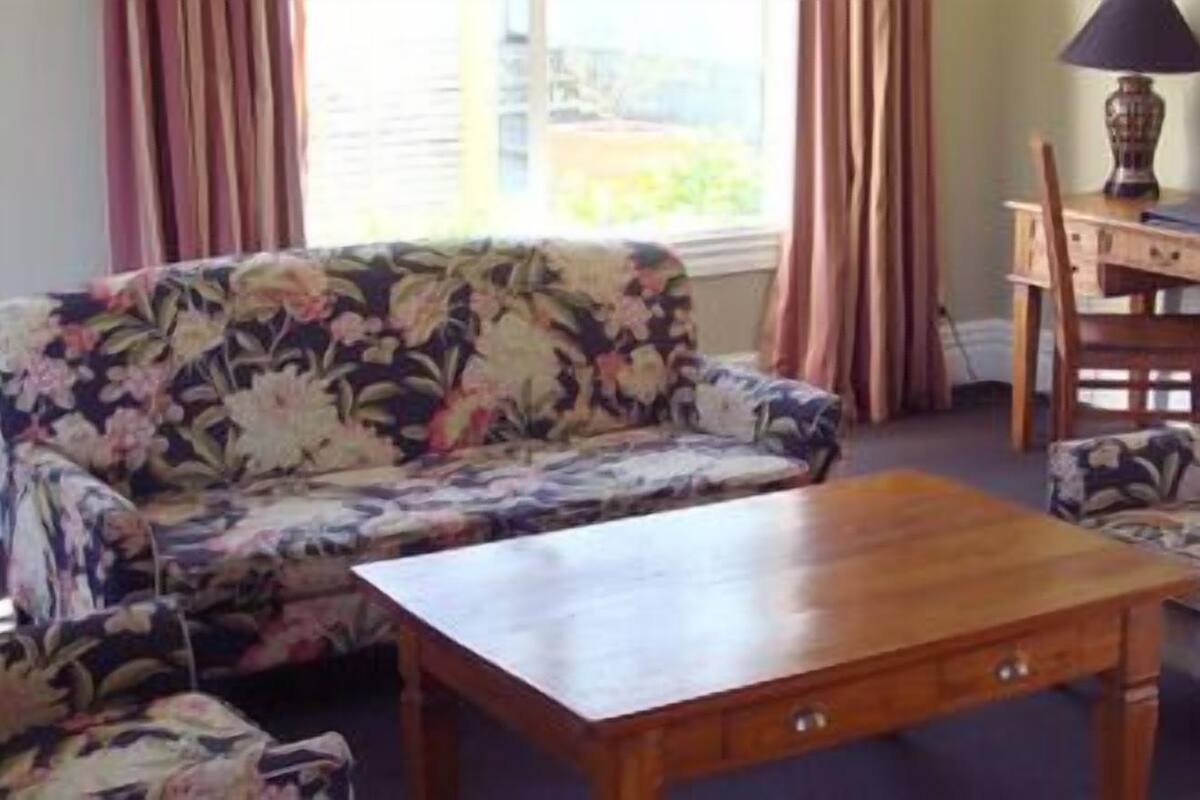 A living room with a floral sofa, a wooden coffee table, and a matching armchair. There is a desk with a lamp and a picture frame.
