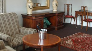 A living room with a striped armchair, a wooden coffee table with a metal pitcher, a red patterned rug, a wooden cabinet, and a mirror on the wall. - The White Swan Country Hotel (Greytown)