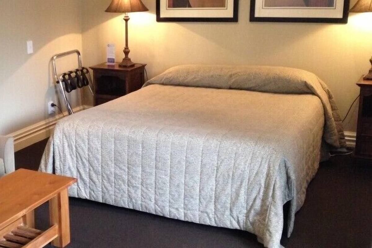 A hotel room with a bed, a sofa, a small wooden table, a nightstand with a lamp, and a wall-mounted coat rack.