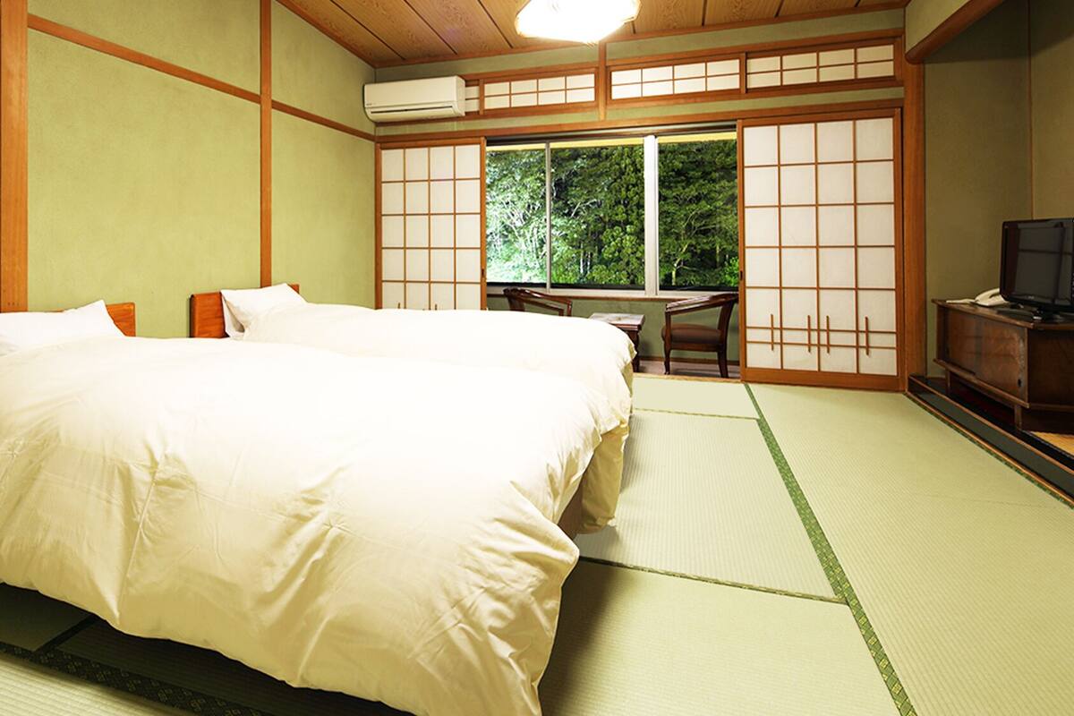 A traditional Japanese-style room with tatami flooring, a large bed with white bedding, a desk with a chair, and a sliding door leading to a view of greenery.