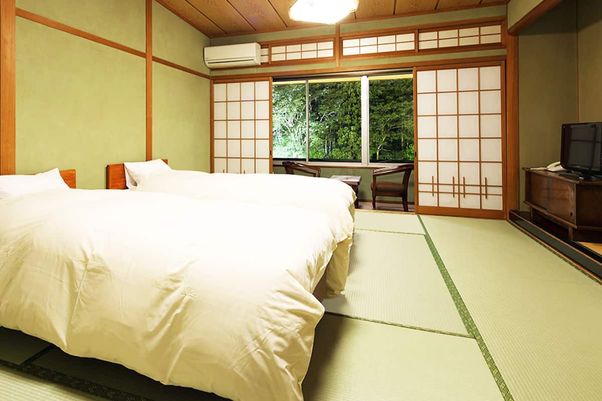 A traditional Japanese-style room with tatami flooring, a large bed with white bedding, a desk with a chair, and a sliding door leading to a view of greenery.