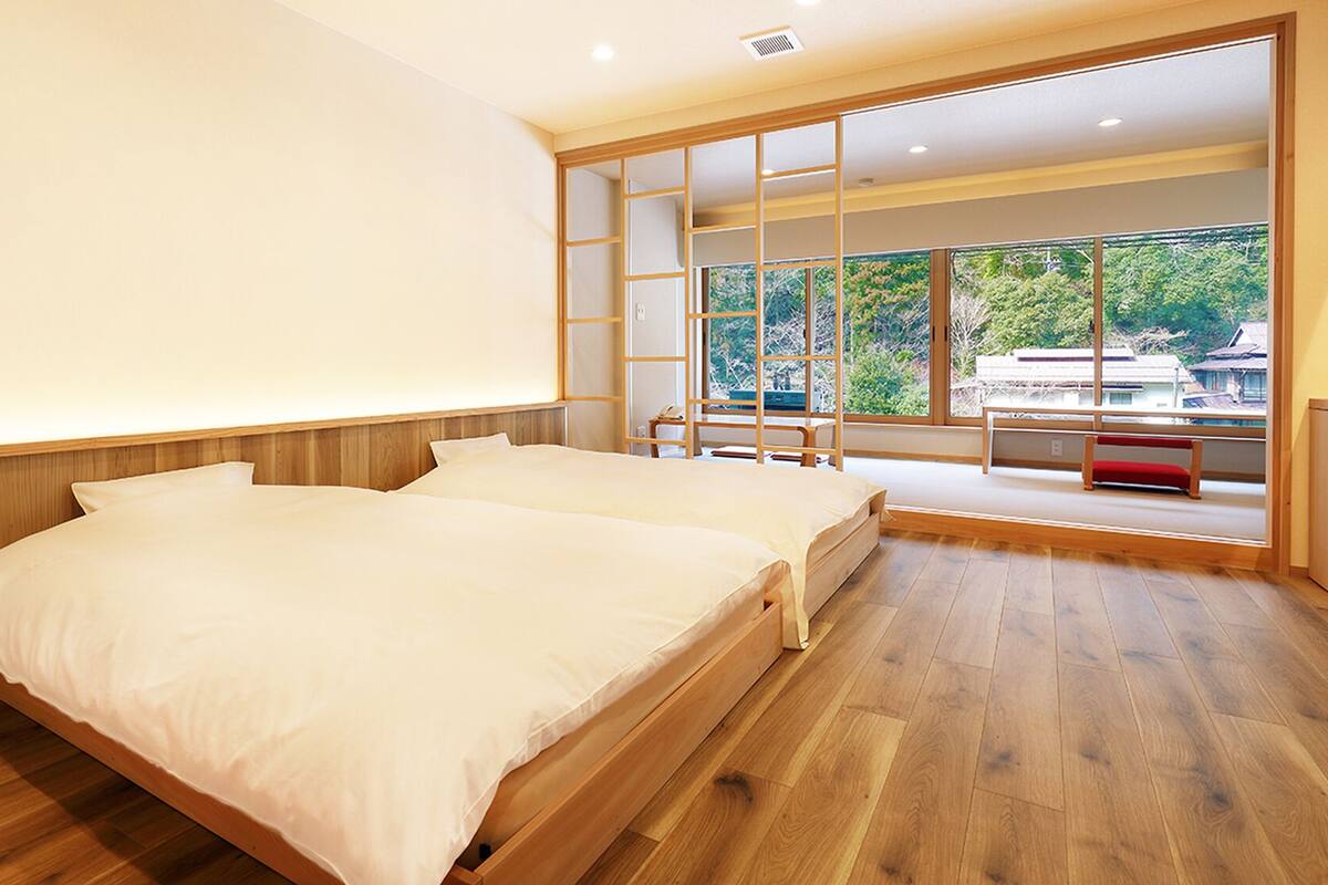 A bedroom with two beds, a sliding glass door leading to a balcony, and a view of greenery outside.