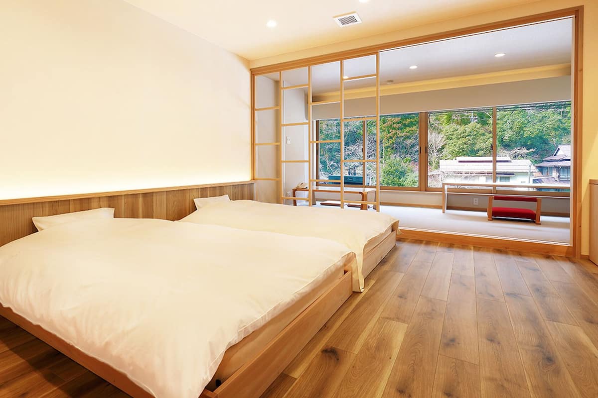 A bedroom with two beds, a sliding glass door leading to a balcony, and a view of greenery outside.