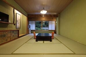Ein Raum mit Tatami-Matten, einem niedrigen Tisch, Stühlen, einem Fernseher an der Wand und Blick ins Grüne.