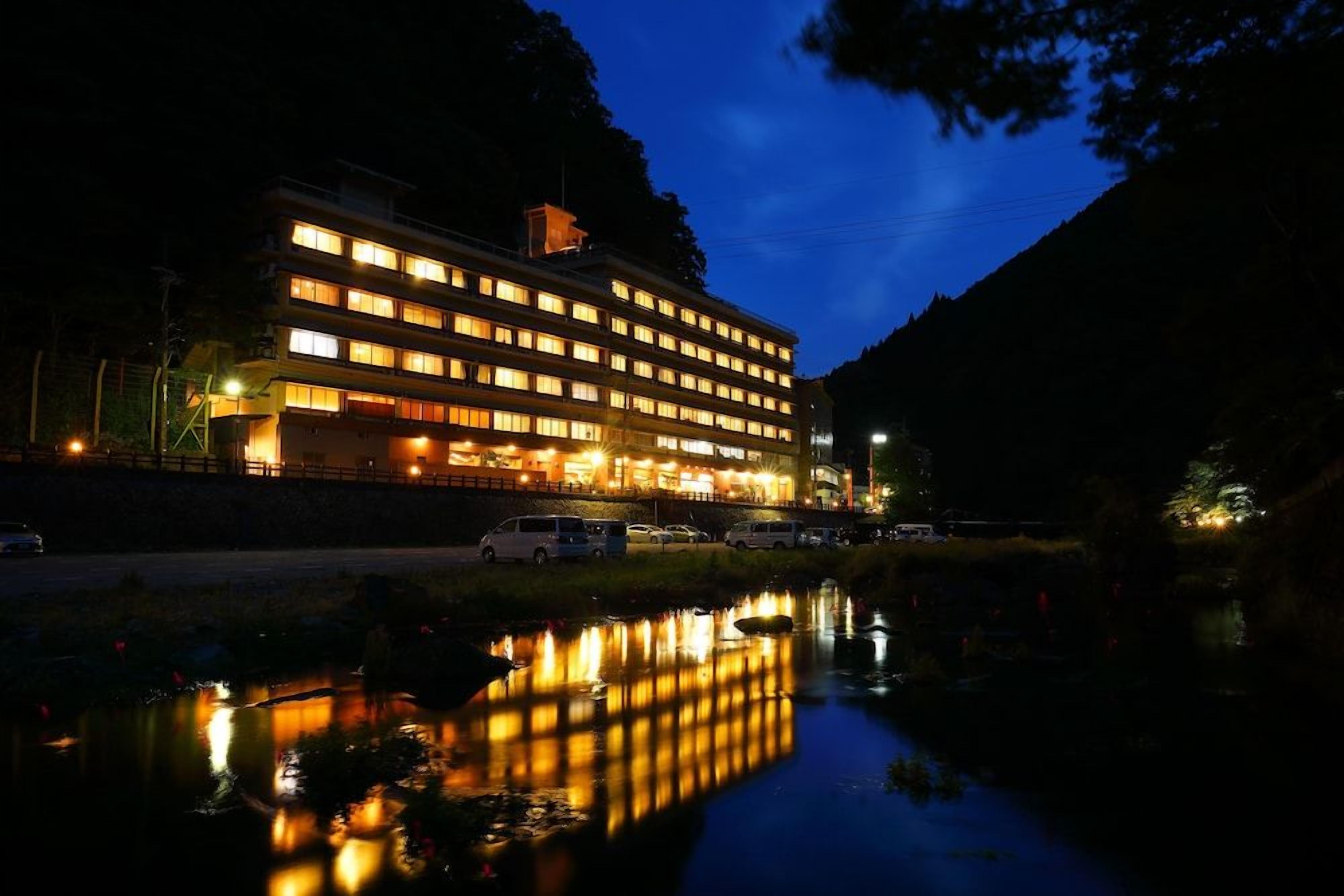 A multi-story hotel with a reflection in a body of water, surrounded by trees and a mountain in the background.