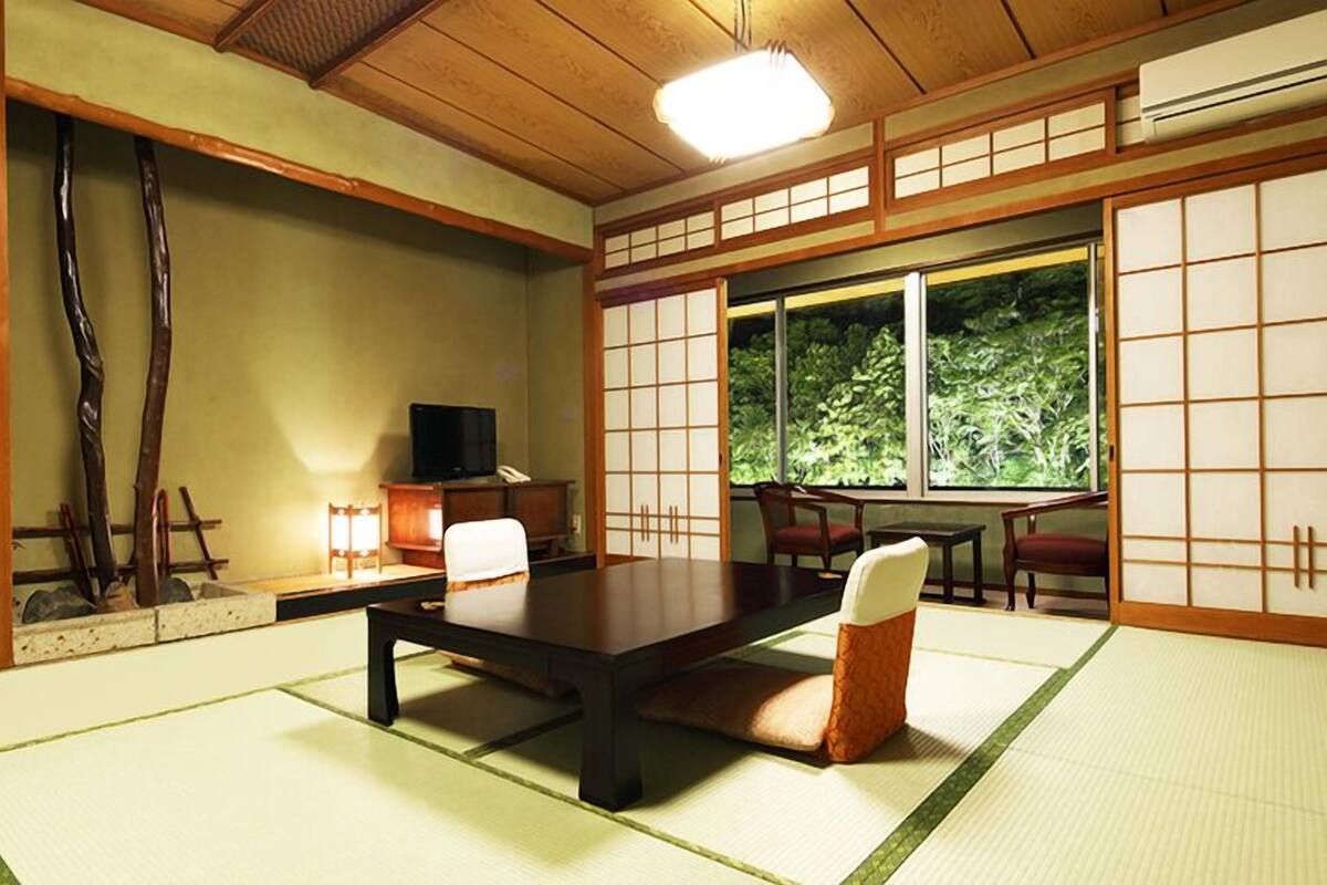 A traditional Japanese room with tatami flooring, a low table, chairs, a television, and a view of greenery through large windows.