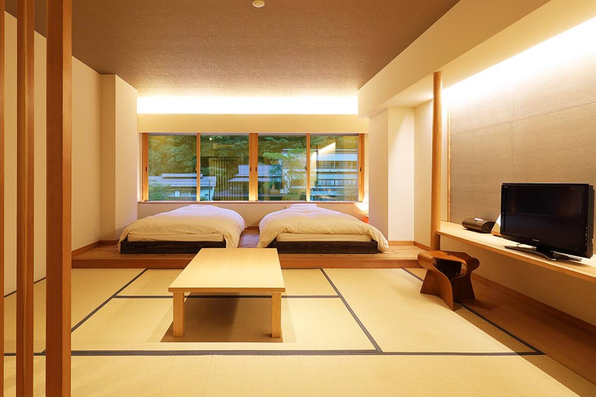 A traditional Japanese room with tatami flooring, two beds, a TV, and a view of a building through large windows.