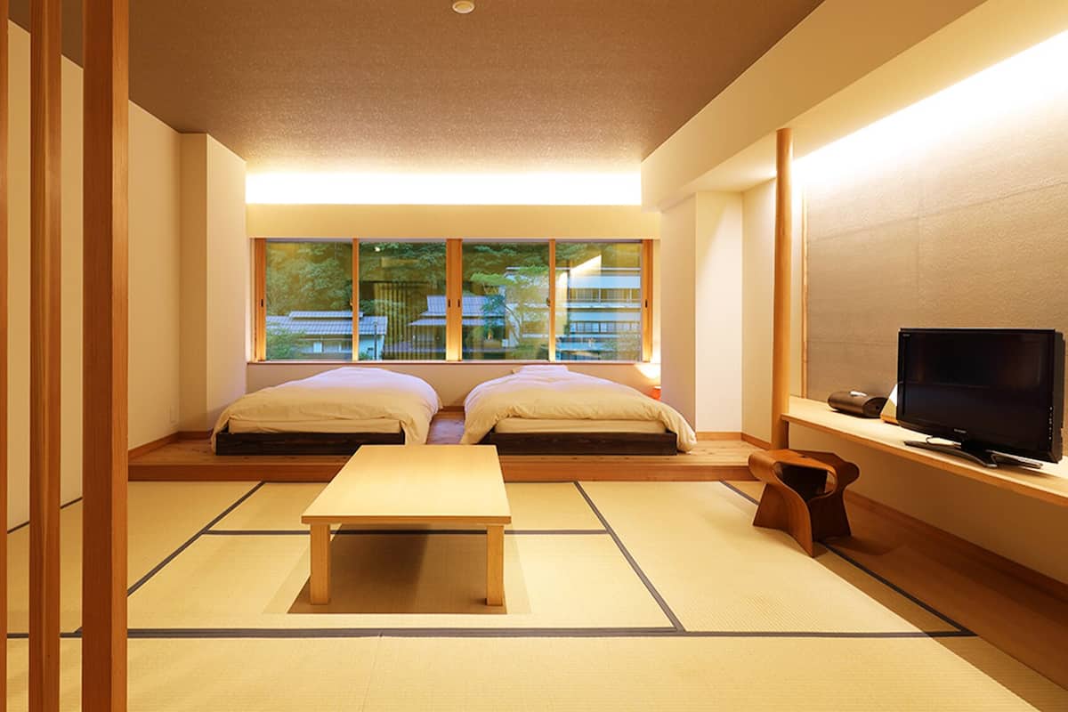 A traditional Japanese room with tatami flooring, two beds, a TV, and a view of a building through large windows.