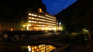 A multi-story hotel with illuminated windows, a parking area, and a body of water reflecting the lights. - Ryokan Yubara International Tourist HOT (MANIWA SHI)