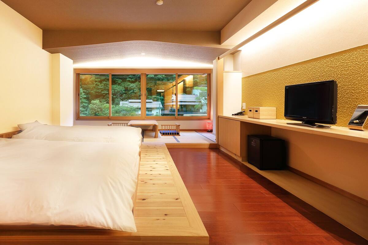 A modern hotel room with a large bed, a desk with a flat-screen TV, and a view of greenery through large windows.