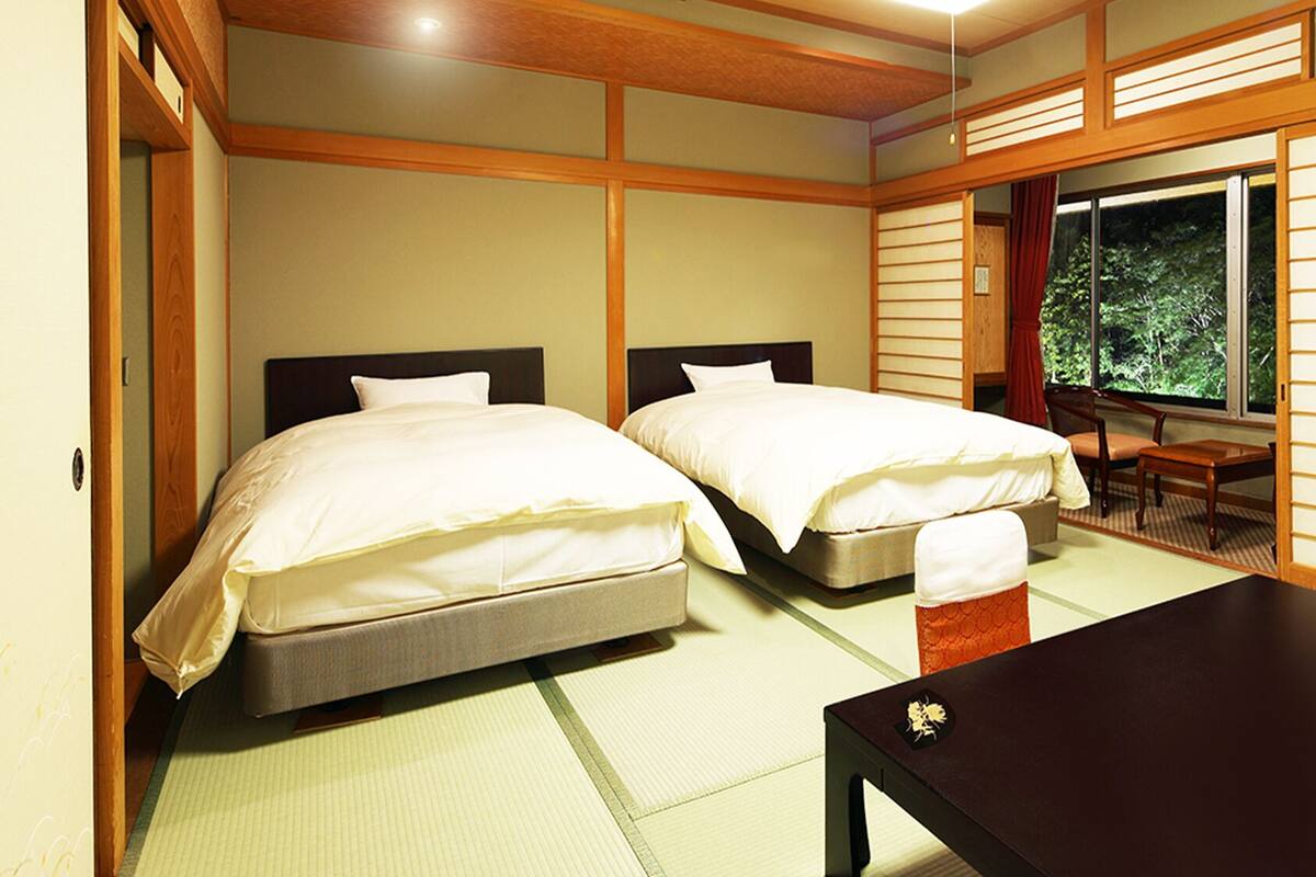 A room with two beds, a small table, and a view of greenery through a window.