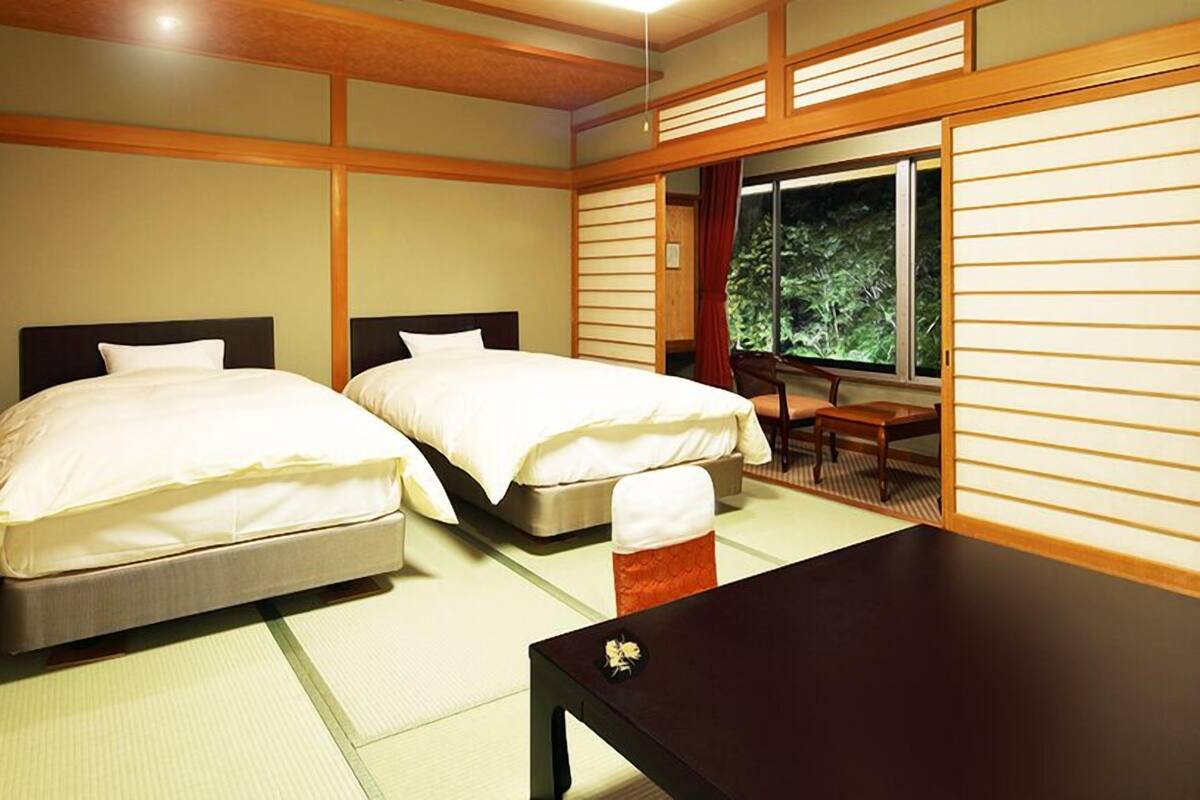 A room with two beds, a small table, and a view of greenery through a window.