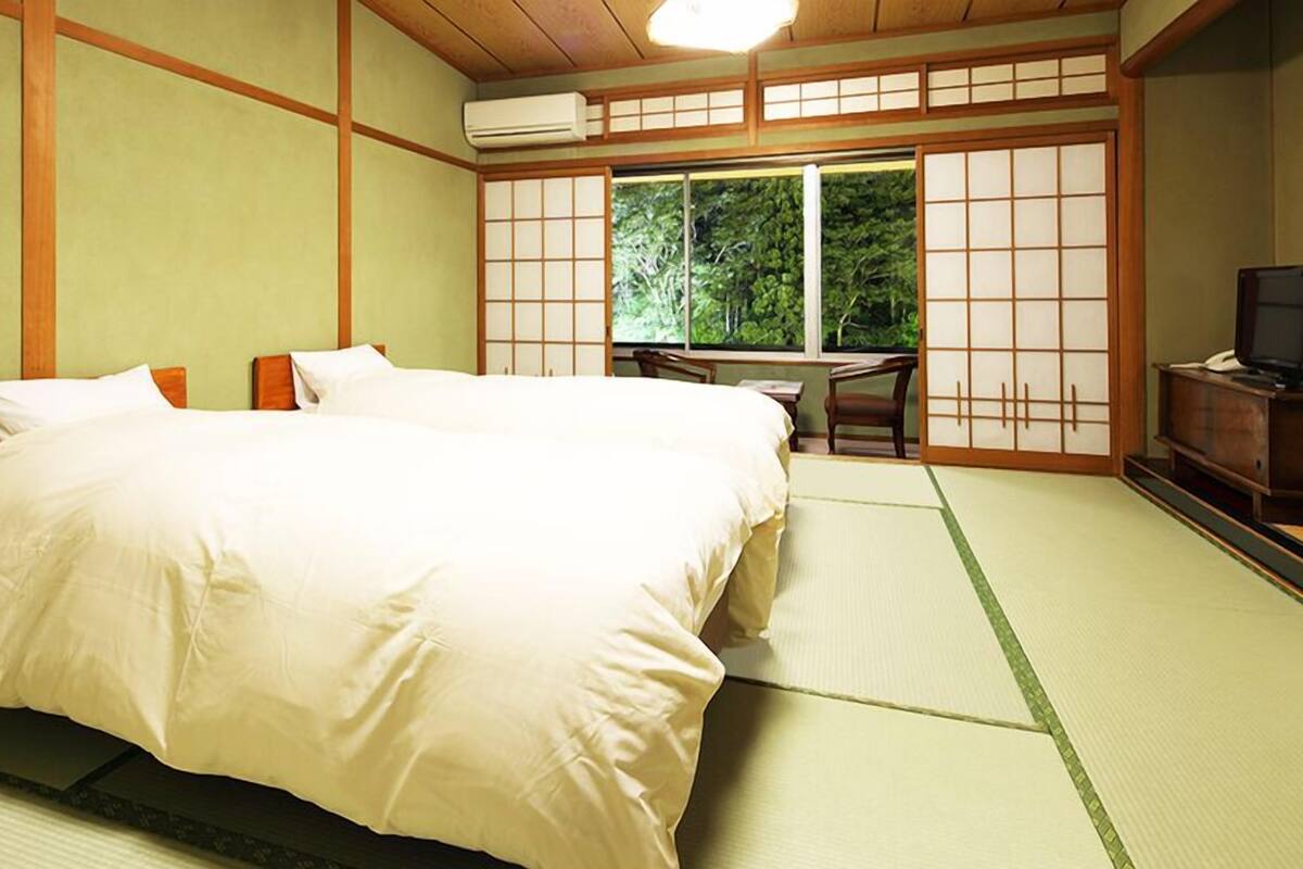 A traditional Japanese-style room with tatami flooring, a bed with white bedding, a desk with a chair, and a sliding door leading to a balcony with a view of greenery.