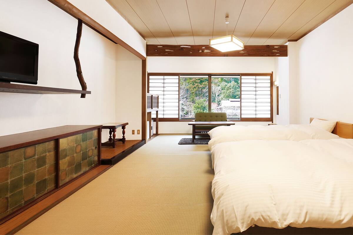 A hotel room with a bed, a TV, a desk, and a view of trees through the window.