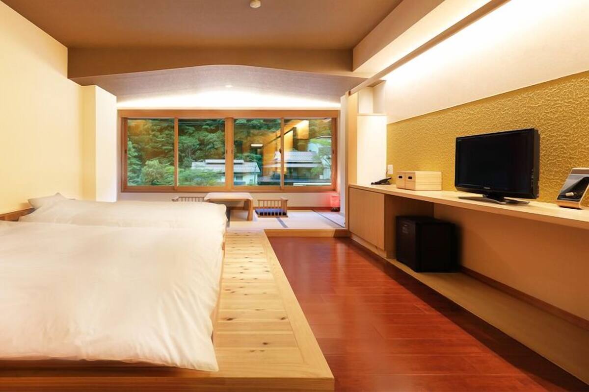 A modern hotel room with a large bed, a desk with a computer, a TV, and a view of greenery outside.