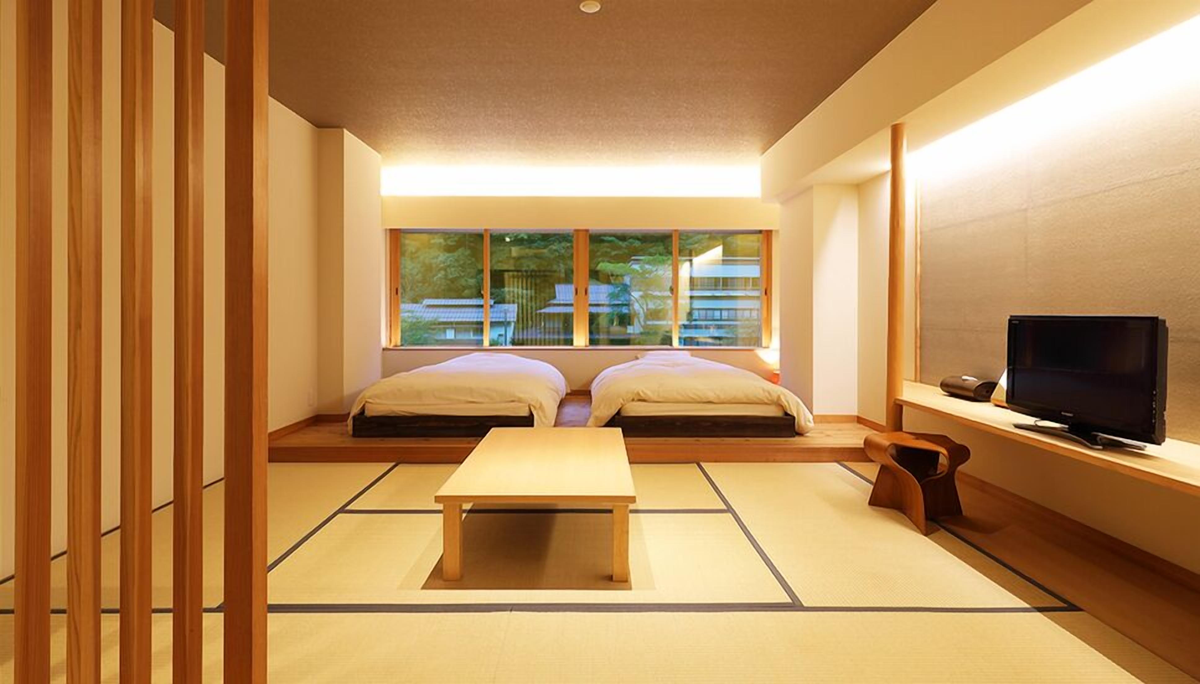 A traditional Japanese room with tatami flooring, a low wooden table, two beds, a TV, and a view of buildings outside.