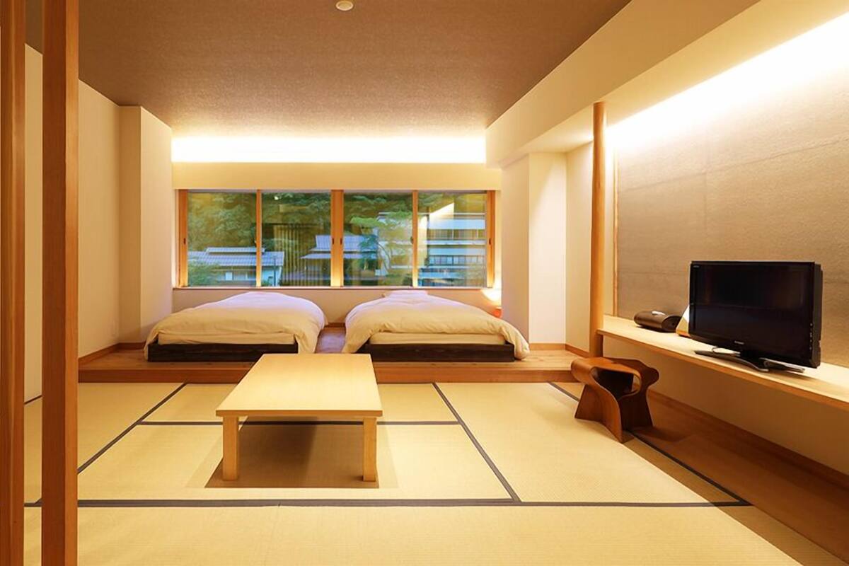 A traditional Japanese room with tatami flooring, a low wooden table, two beds, a TV, and a view of buildings outside.