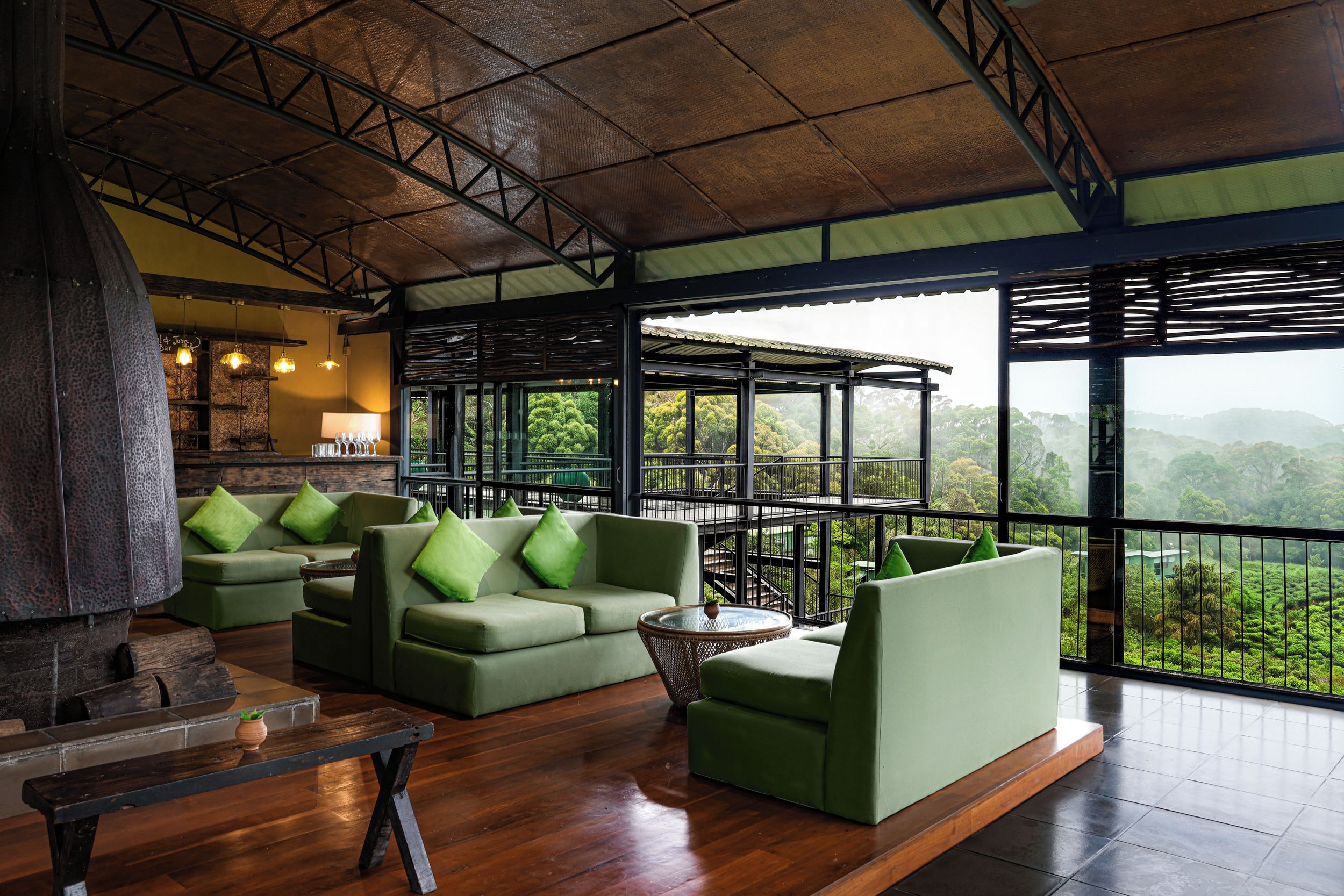 A spacious lounge area with a wooden floor, green sofas with cushions, a round glass table, and a view of lush greenery through large windows.