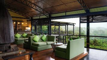 A spacious lounge area with a wooden floor, green sofas with cushions, a round glass table, and a view of lush greenery through large windows.