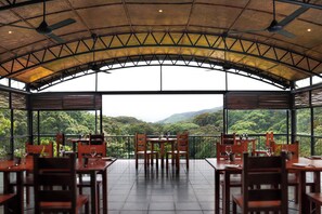 Un área de comedor al aire libre con mesas y sillas, amplios ventanales con vistas a un paisaje boscoso.