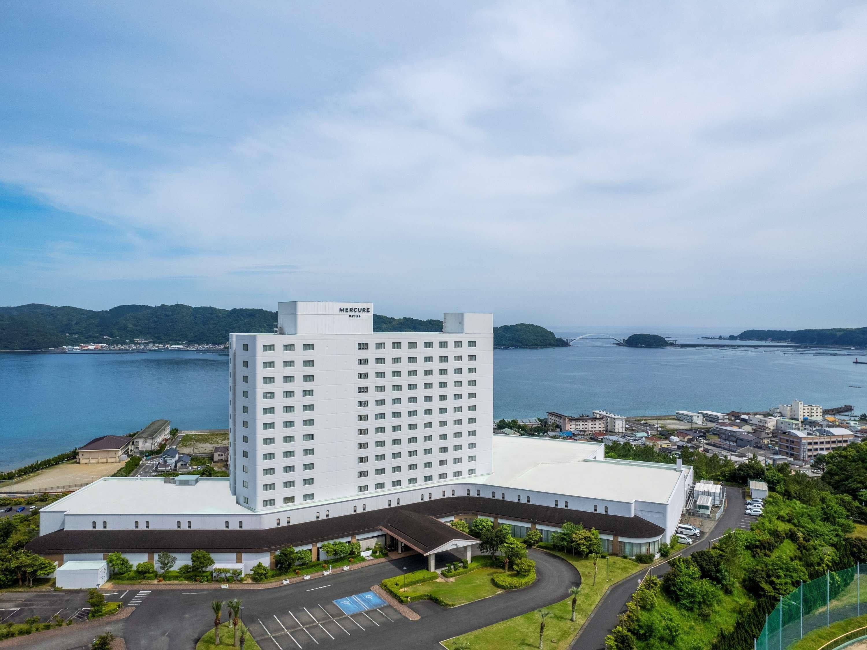 A large, modern hotel with a prominent 'Mercure' sign, surrounded by a parking area and greenery, with a view of a body of water and distant hills.