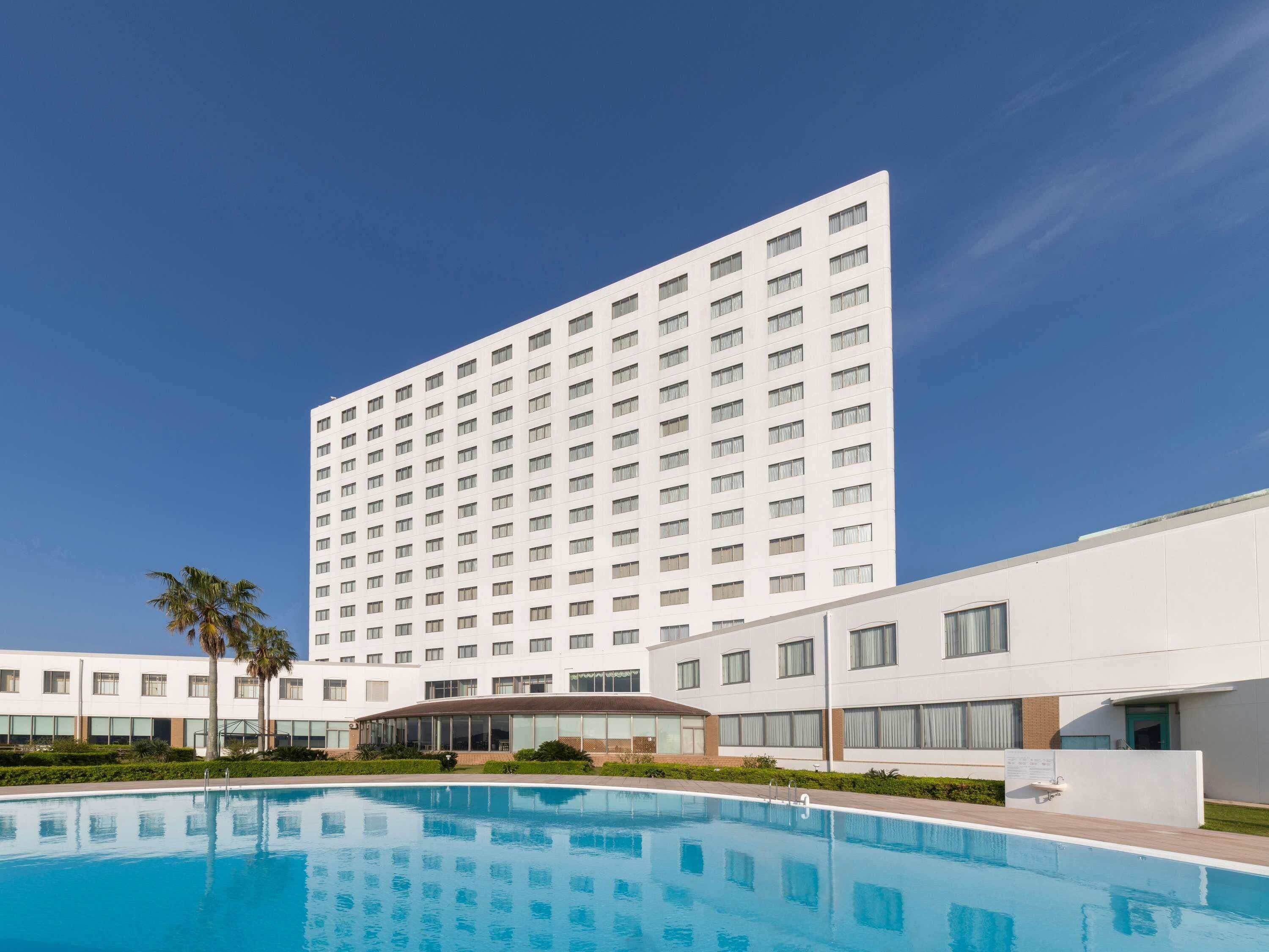 Un hotel moderno con una piscina grande, varios pisos y un cielo azul despejado.