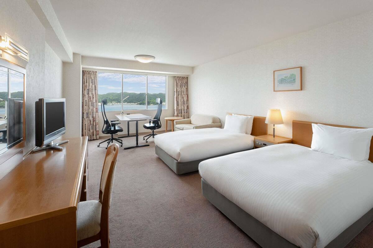 Ein Hotelzimmer mit zwei Betten, einem Schreibtisch mit Computer, einem Stuhl, einem kleinen Tisch und Blick aufs Meer.