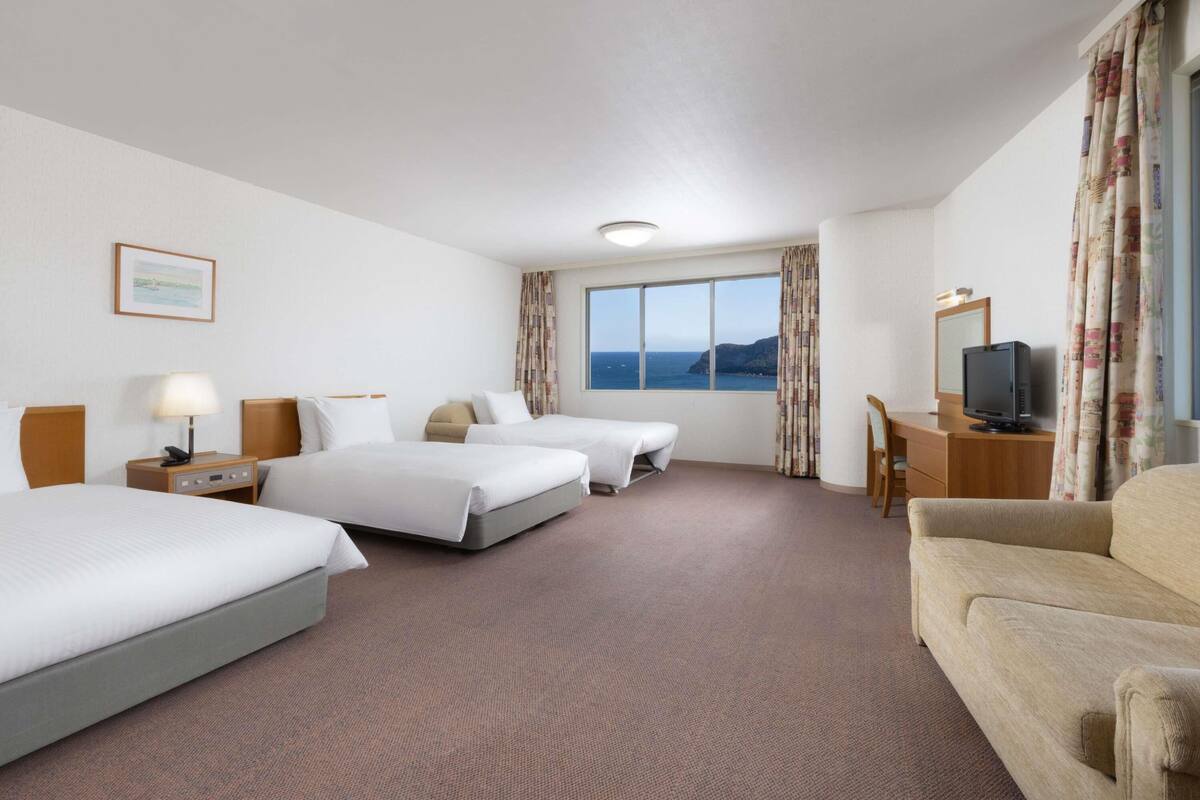 Ein Hotelzimmer mit zwei Betten, einem Schreibtisch mit Fernseher, einem Sessel und Meerblick.