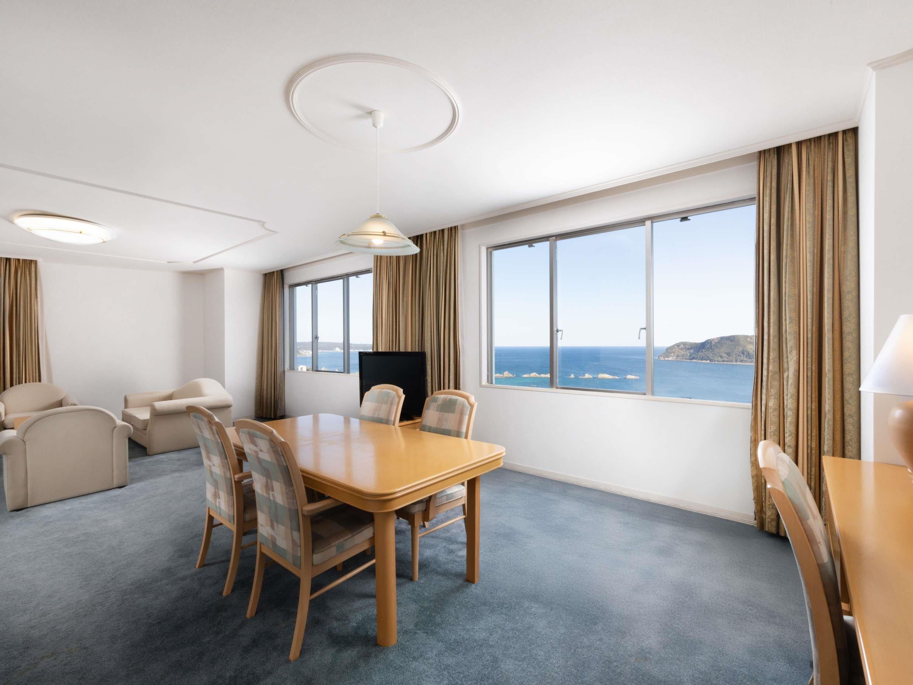 Habitación de hotel con un amplio ventanal que ofrece vistas al mar, una mesa de comedor de madera con sillas, un televisor de pantalla plana y un sofá.