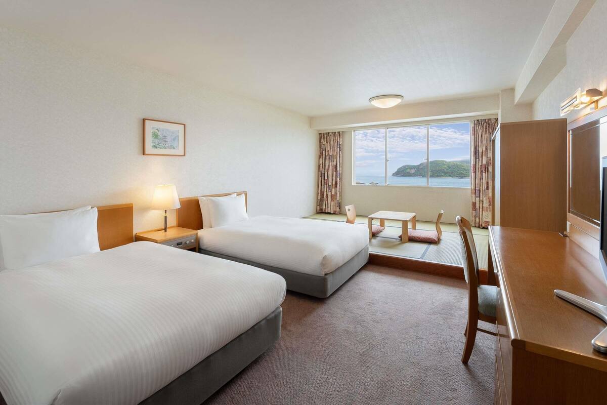 Ein Hotelzimmer mit zwei Betten, einem Schreibtisch, einem Sessel, einem Fernseher und einem Fenster mit Meerblick.