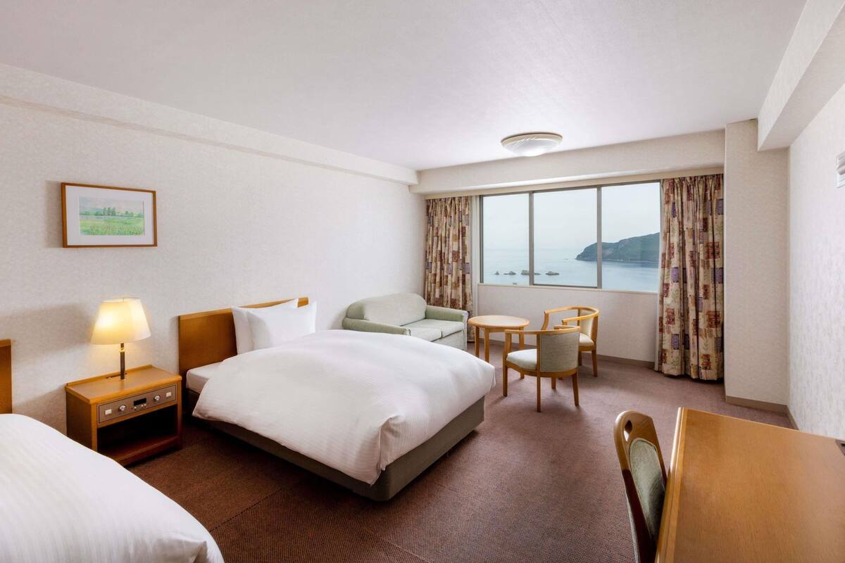 Ein Hotelzimmer mit einem Bett, zwei Stühlen, einem kleinen Tisch, einem Nachttisch, einer Lampe, einem Fenster mit Meerblick und einem gerahmten Bild an der Wand.