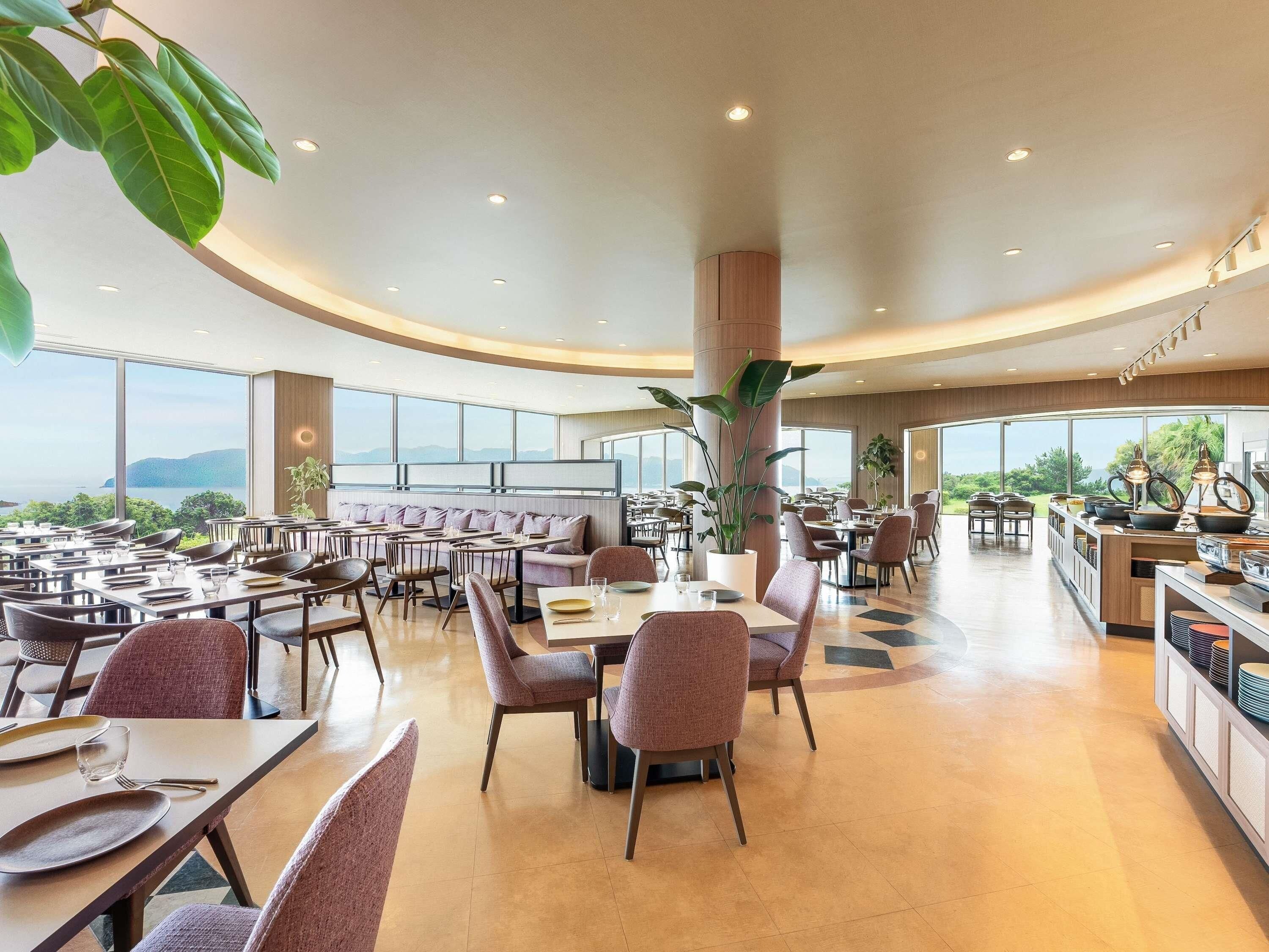 A spacious restaurant with high ceilings, large windows, and a central column with plants. Tables and chairs are arranged for dining.