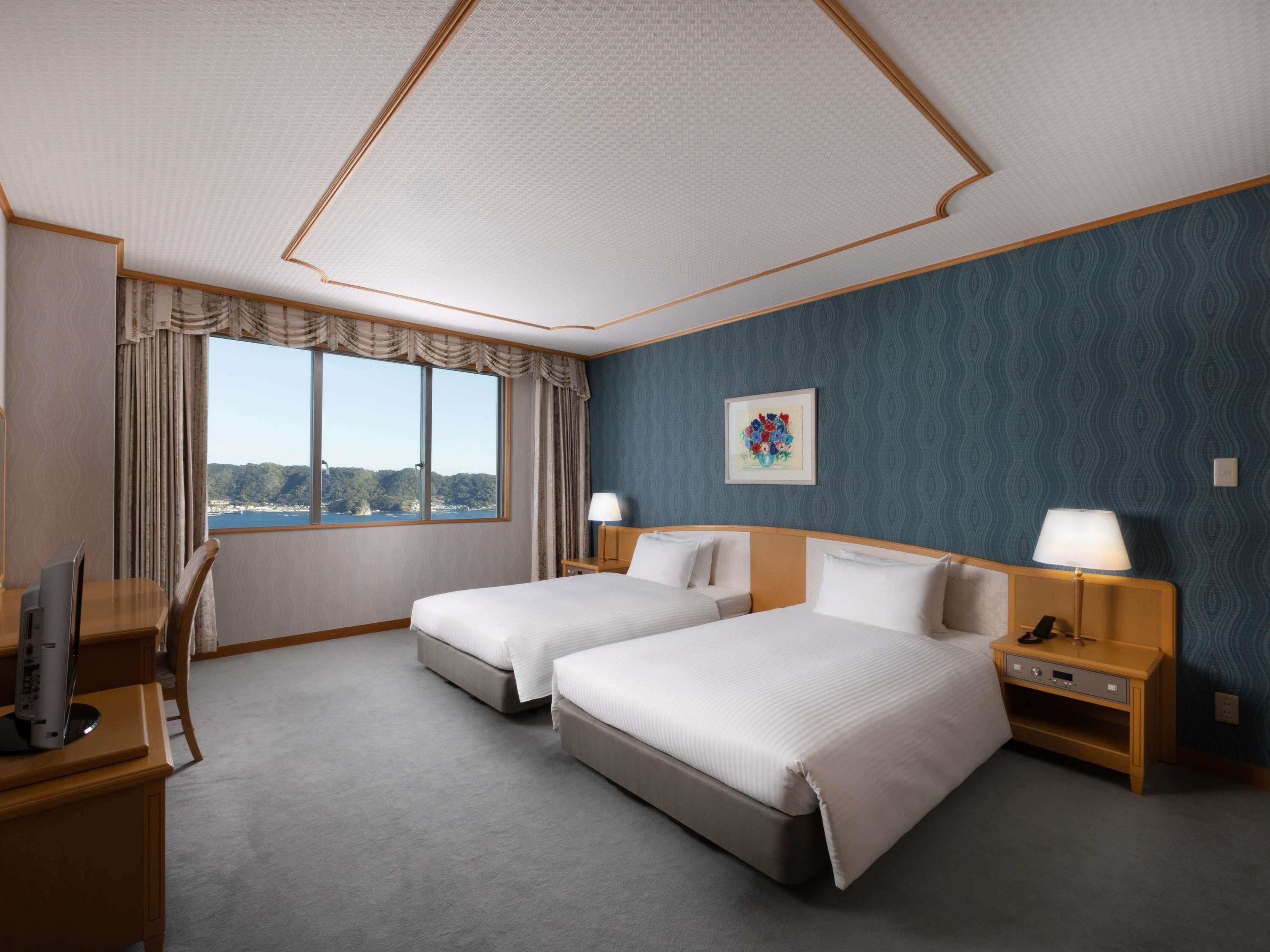 A hotel room with two beds, a desk with a computer, a window with a view, and a painting on the wall.