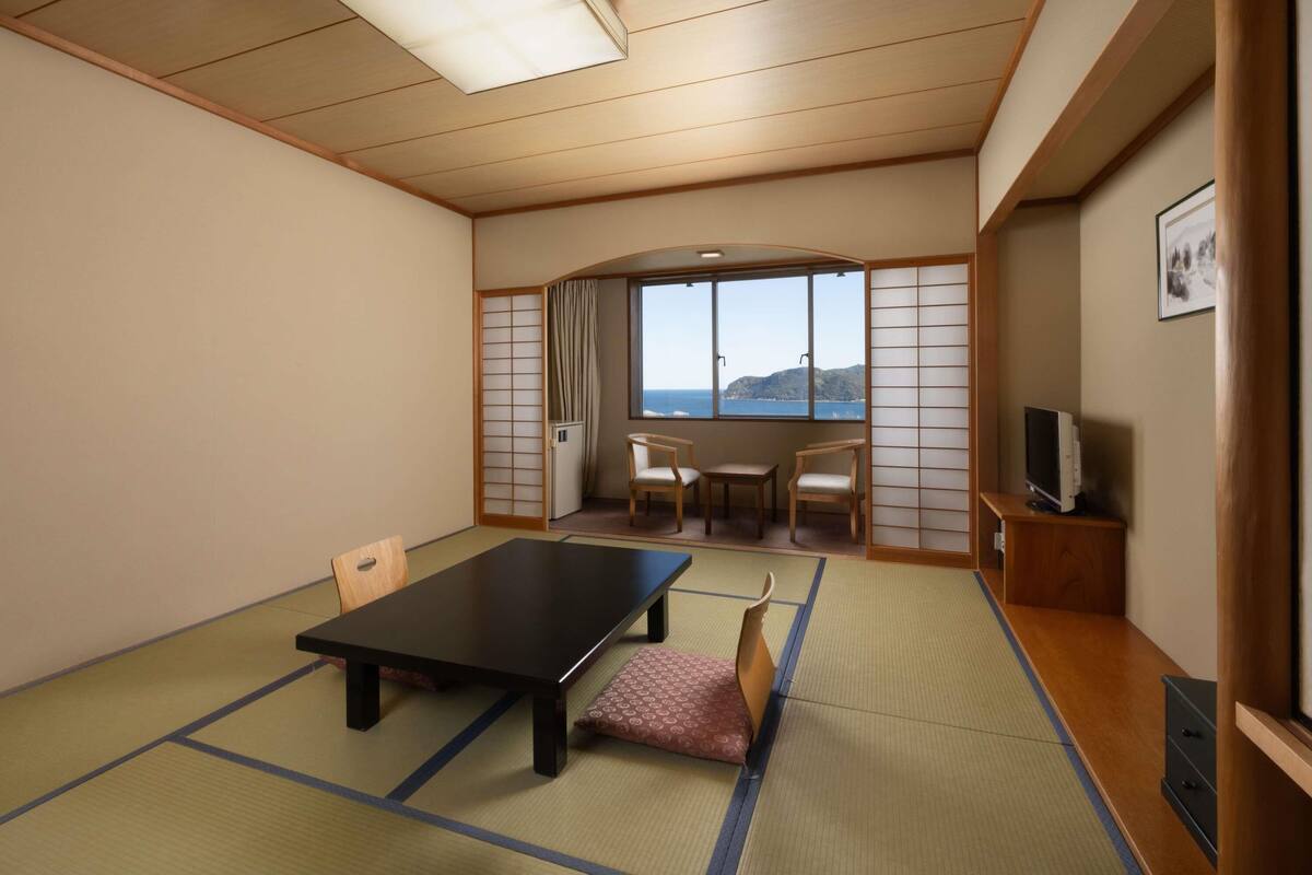 Ein traditioneller japanischer Raum mit Tatami-Böden, einem niedrigen Tisch, Stühlen, einem Fernseher und Blick aufs Meer.