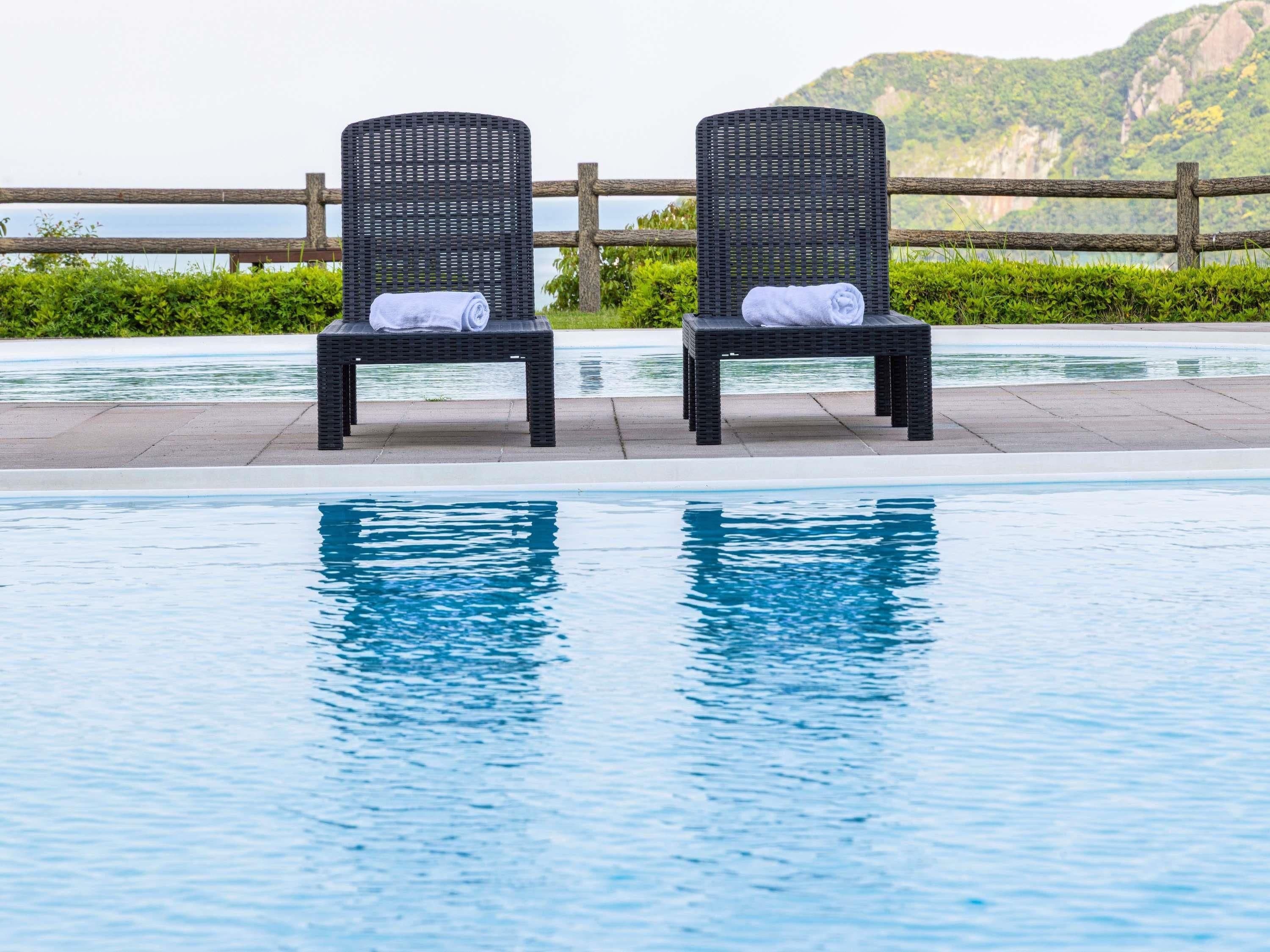 Two wicker lounge chairs with towels, a swimming pool, and a wooden fence with a view of a mountainous landscape.