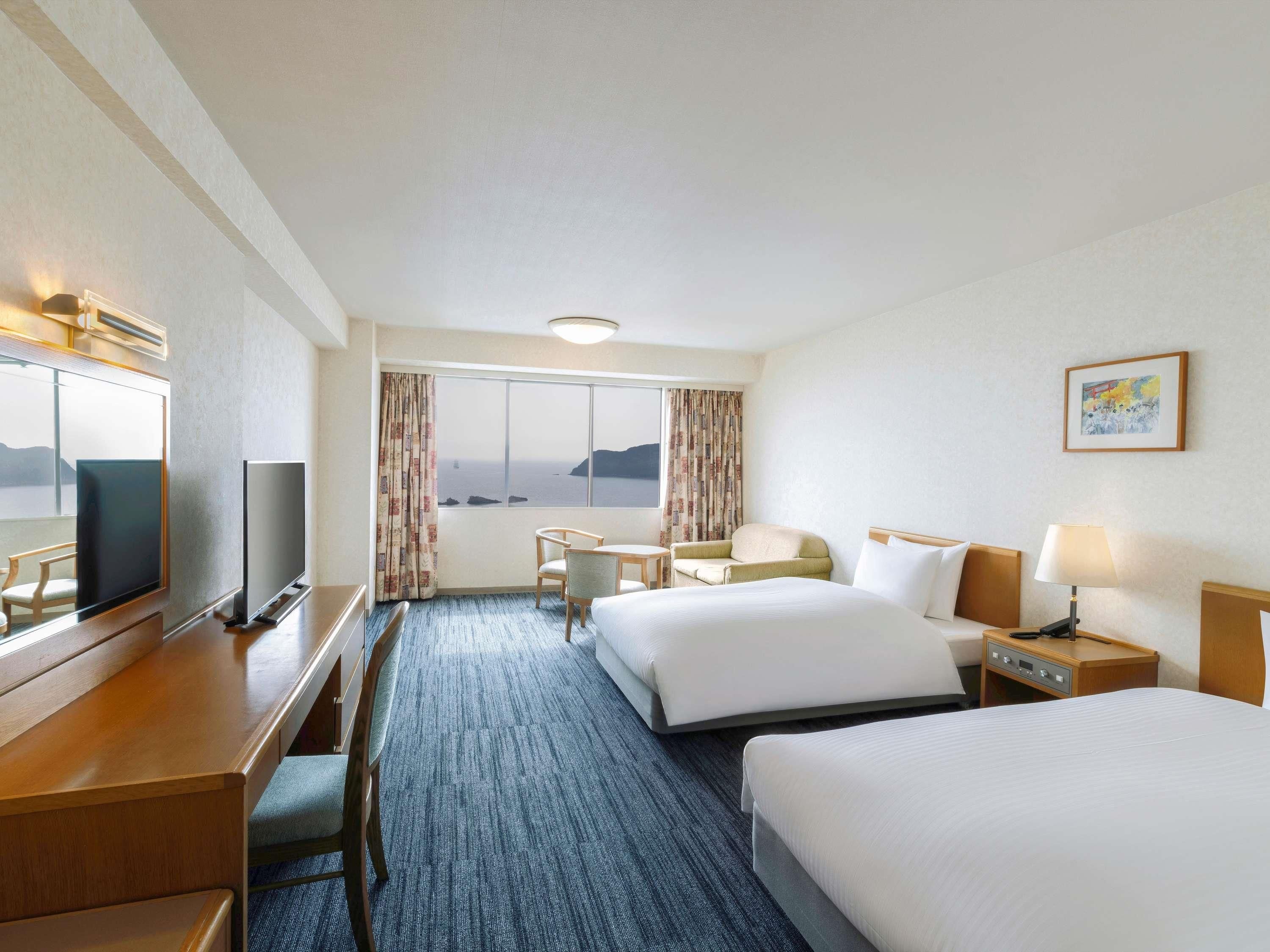 Une chambre d’hôtel avec deux lits, un bureau avec une télévision, une chaise, une petite table et une fenêtre donnant sur un paysage agréable.
