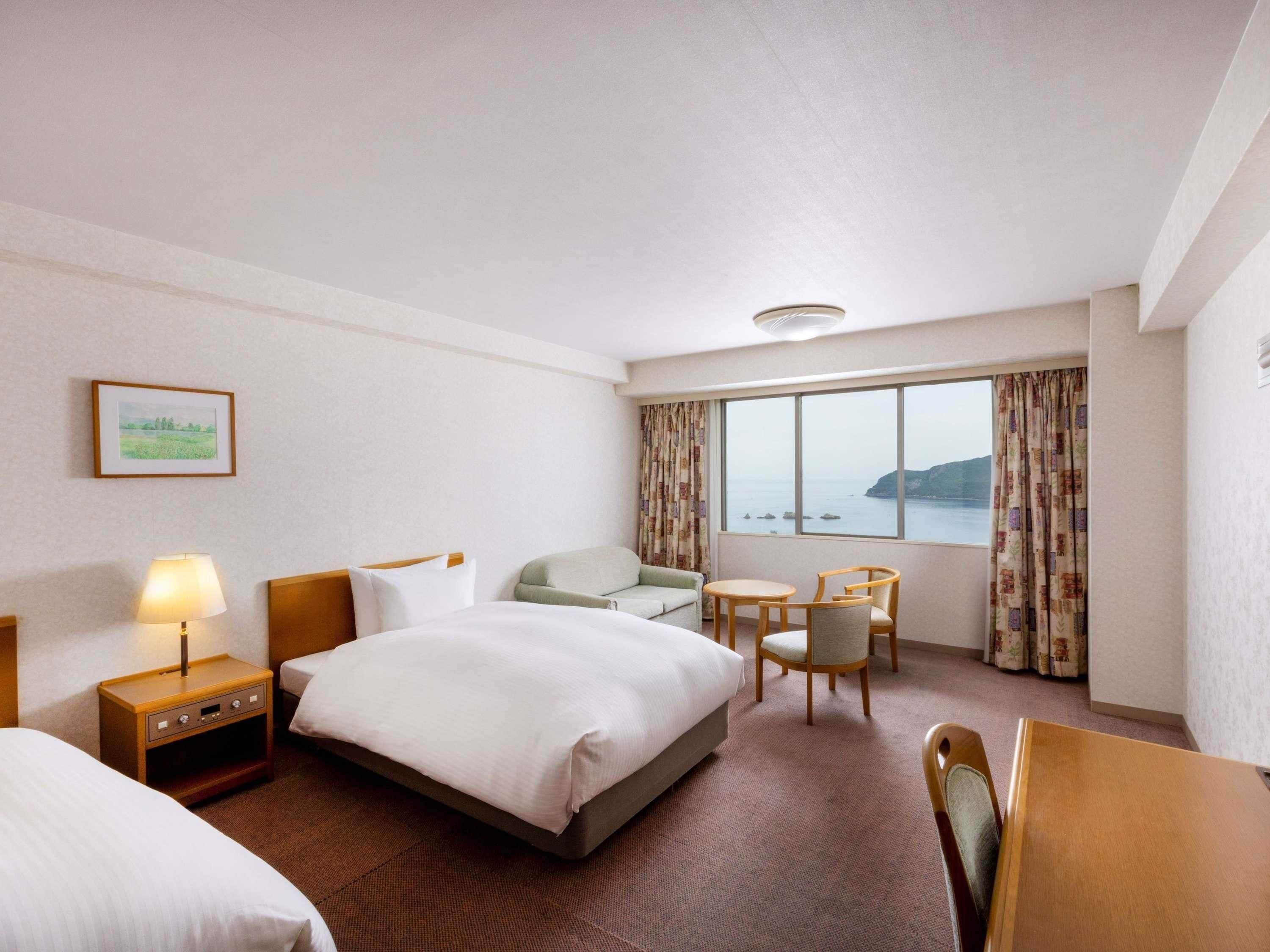 A hotel room with a bed, two chairs, a small table, a nightstand, a lamp, a window with a view of the sea, and a framed picture on the wall.