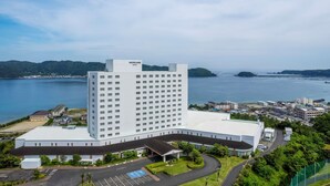 Un hotel grande y moderno con un llamativo letrero de 'Mercure', rodeado de un área de estacionamiento y vegetación, con vistas a una masa de agua y colinas distantes.