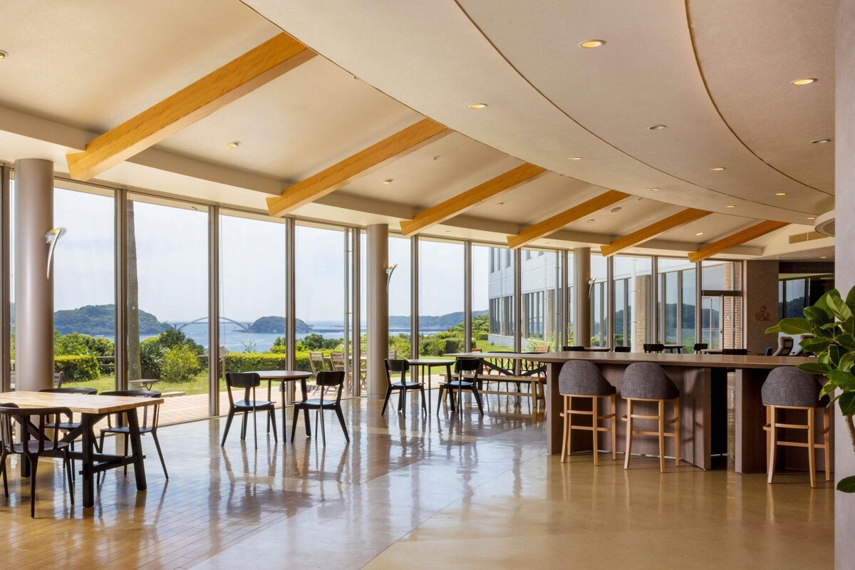 A spacious indoor area with large windows offering a view of a bridge and greenery. There are tables and chairs arranged for dining or meetings.