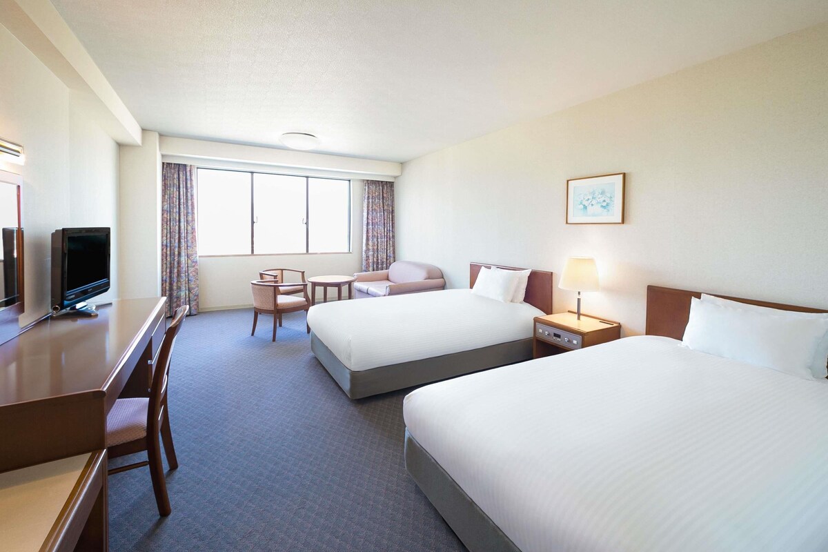 A hotel room with two beds, a desk with a television, a chair, a nightstand with a lamp, and a window with curtains.