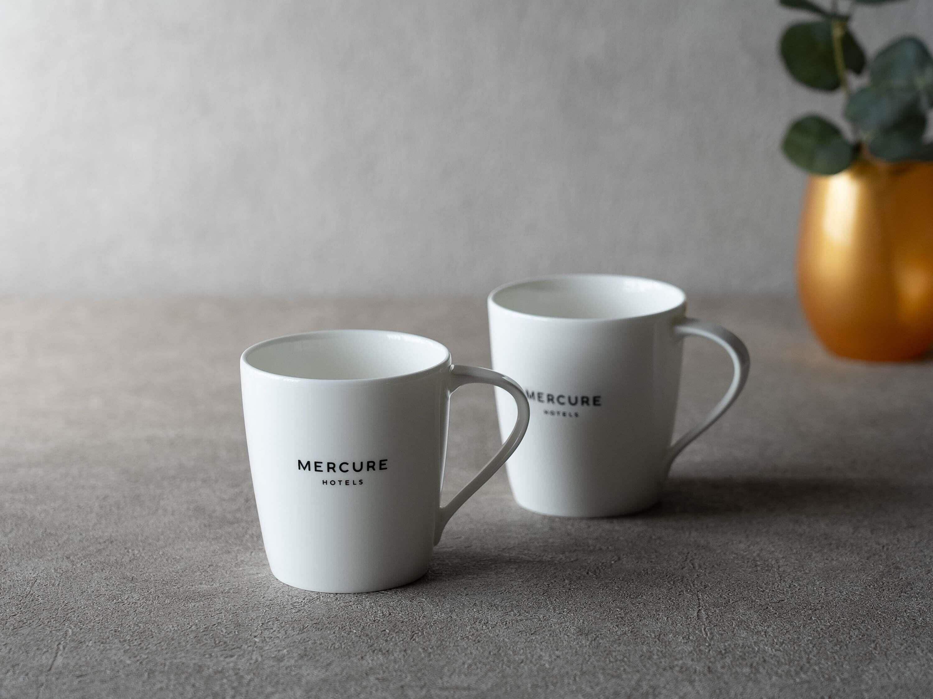 Two white mugs with 'MERCURE HOTELS' text, placed on a textured surface with a blurred background.