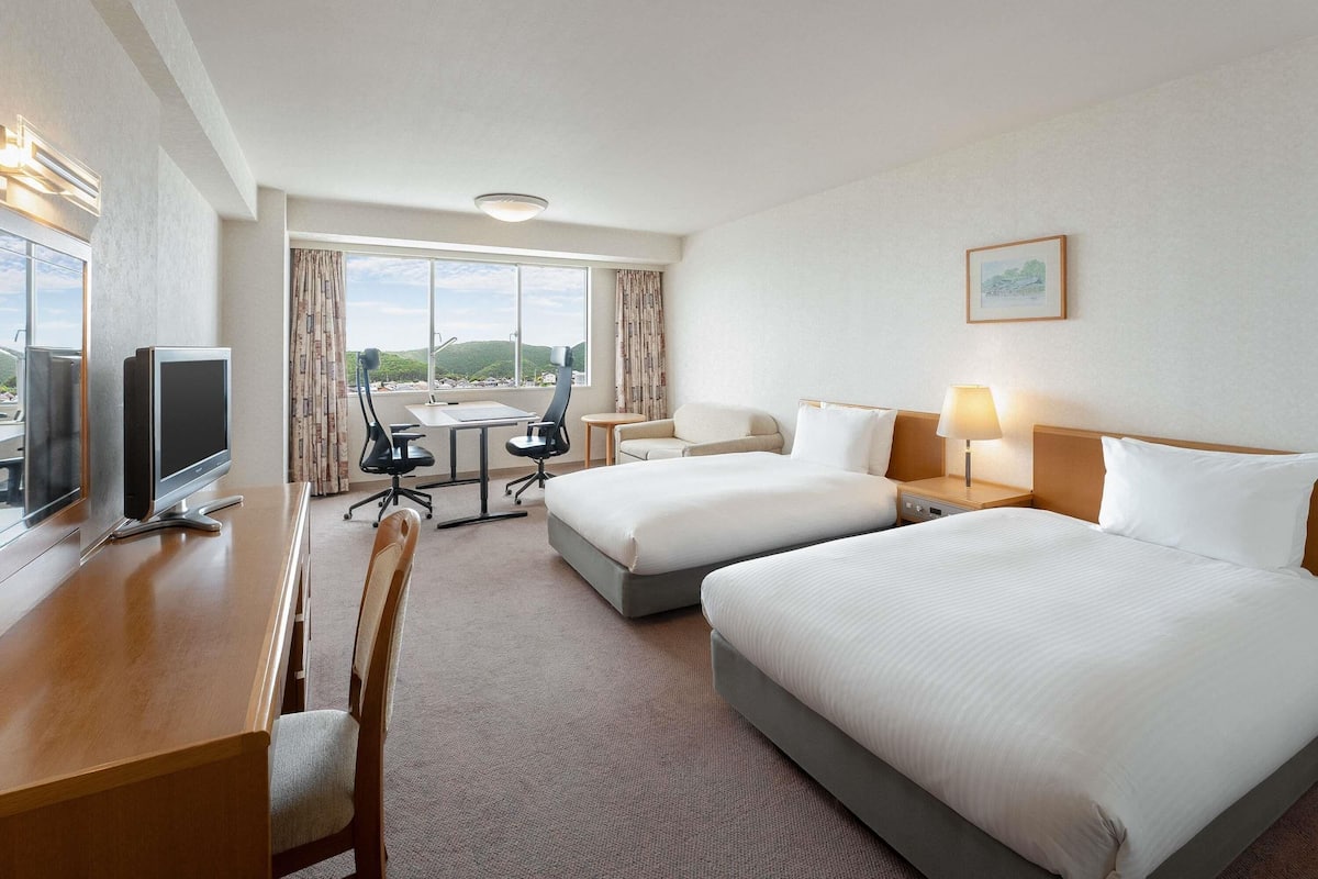 A hotel room with two beds, a desk with a computer, a chair, a small table, and a window with a view of hills.