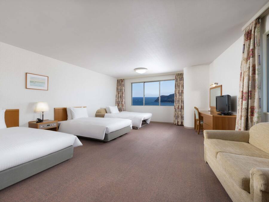 A hotel room with two beds, a desk with a television, a chair, and a view of the sea.