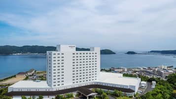 Un hotel grande y moderno con un llamativo letrero de 'Mercure', rodeado de un área de estacionamiento y vegetación, con vistas a una masa de agua y colinas distantes.