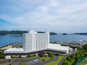 A large, modern hotel with a prominent 'Mercure' sign, surrounded by a parking area and greenery, with a view of a body of water and distant hills. - Mercure Wakayama Kushimoto Resort & Spa (Kushimoto)