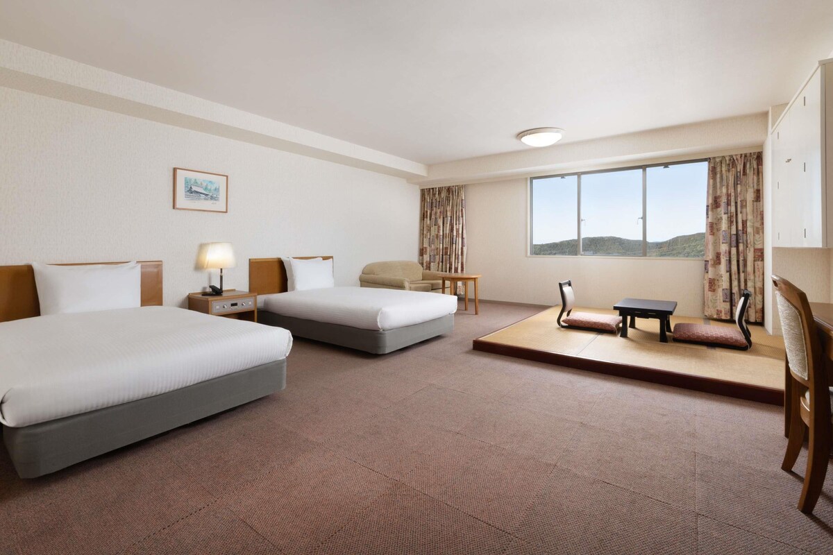A hotel room with two beds, a desk, a chair, a small table, a window with a view, and a painting on the wall.