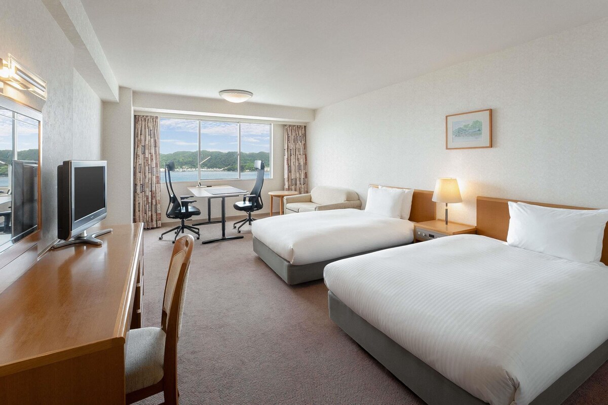 A hotel room with two beds, a desk with a computer, a chair, a small table, and a view of the sea.