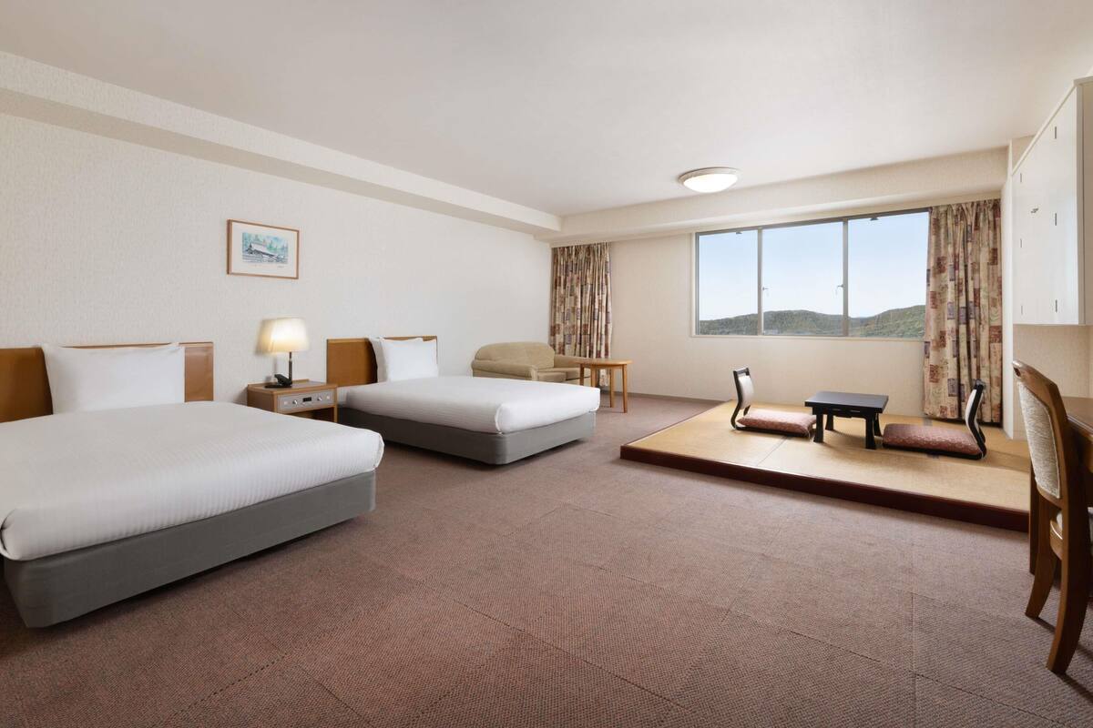 Ein Hotelzimmer mit zwei Betten, einem Schreibtisch, einem Sessel, einem kleinen Tisch, einem Fenster mit Blick und einem Bild an der Wand.