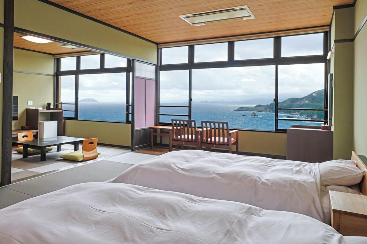 A room with two beds, a desk, a chair, and large windows offering a view of the sea.