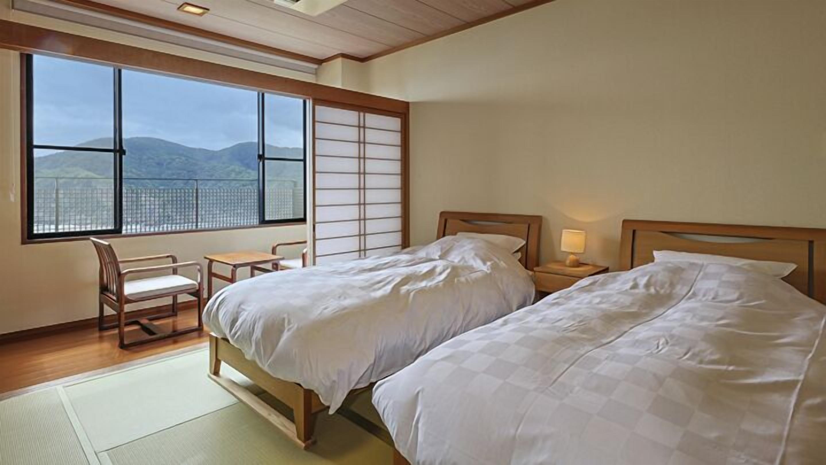 A bedroom with two beds, a desk, and a chair, with a view of mountains through large windows.